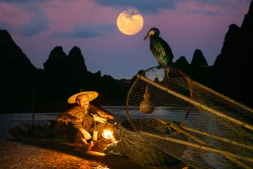 Photo sur Plexiglas Guilin Traditional Chinese Fisherman taking a break on bamboo raft with two cormorants illuminated by his flaming (petrol) lantern on the Li River with full moon rising at behind the Guilin Hills, China
