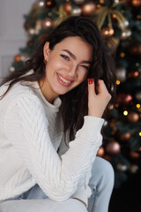 Portrait of pretty brunette woman near New Year Tree. Christmas holyday. Happy girl in cosy sweater sitting on beige sofa, smile. Celebrate New Year Eve. Woman at home relax