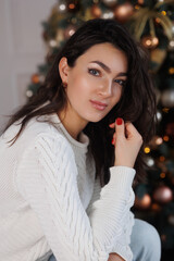 Portrait of pretty brunette woman near New Year Tree. Christmas holyday. Happy girl in cosy sweater sitting on beige sofa, smile. Celebrate New Year Eve. Woman at home relax