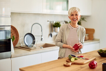 A portrait of a cheerful senior woman standing in the kitchen and preparing fresh vegetables for a salad