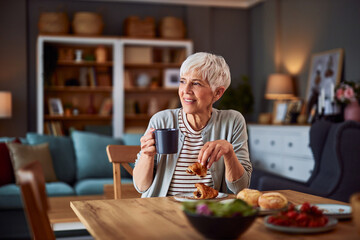 A contented senior adult woman sitting at a dining table and looking away while enjoying her...