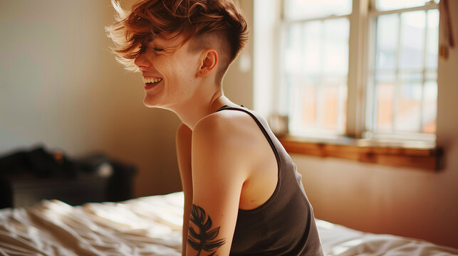 Smiling butch queer woman sitting, sidecut hair, tattoo visible, sunlit room.