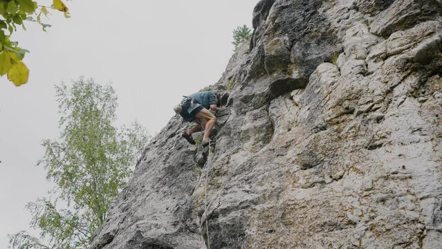 A climber in athletic attire scales a steep limestone cliff, focusing intently on the next move, in a display of strength and agility against the backdrop of a forested landscape. A male climber