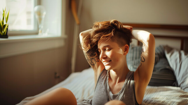 Smiling queer person sitting, stretching, touching hair, tattoo visible, sunlit room.