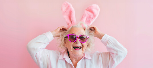 Radiant senior woman with fluffy pink bunny ears laughing heartily, captured against a pastel pink background, exuding Easter cheer