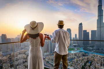 A elegant couple stands on a balcony and enjoys the sunset behind the skyline of Dubai with a drink...