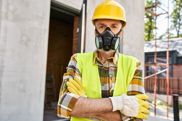 attractive man in safety gloves and helmet wearing dust mask and looking at camera, cottage builder
