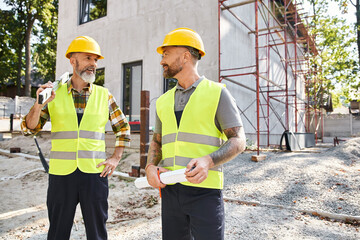 appealing joyous construction workers with level and blueprint looking at each other, builders