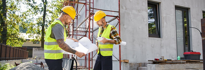 attractive cottage builders in safety vests working on their blueprints on construction site, banner