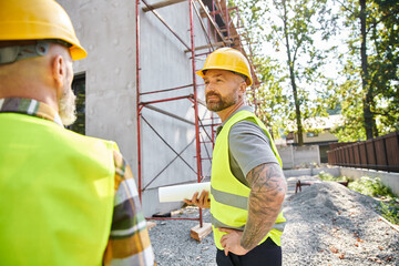 focus on devoted cottage builder with blueprint posing next to his blurred colleague and scaffolding - obrazy, fototapety, plakaty