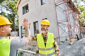 two cheerful cottage builders in safety attires giving high five to each other near scaffolding