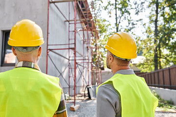 back view of cottage builders in safety helmets looking at scaffolding on construction site - 742714528