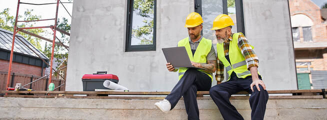 appealing bearded cottage builders in safety helmets sitting on porch and working on laptop, banner