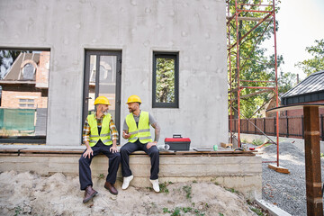 joyful dedicated cottage builders in safety vests sitting on porch and looking at each other