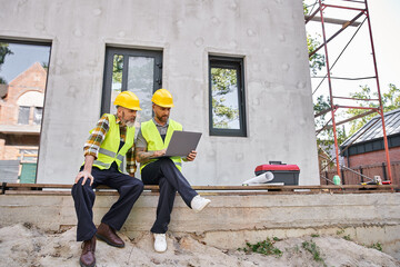 appealing bearded cottage builders in safety helmets sitting on porch and working on laptop