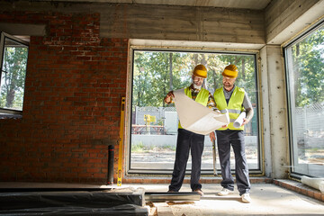 cheerful bearded men in safety helmets looking at blueprint while on construction site, builders - 742715583