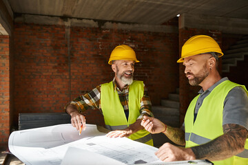 joyous handsome builders in safety vests and helmets looking at blueprint before construction