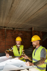 two handsome builders in safety helmets and vests working with blueprint before construction - 742715746