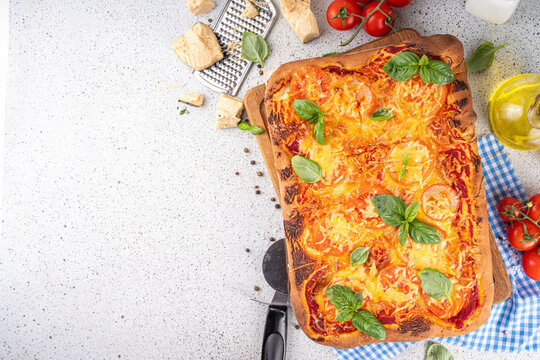 Rectangular homemade margarita pizza with tomatoes and basil, with ingredients and olive oil on white kitchen table background