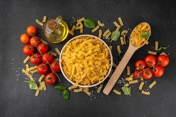 Raw uncooked Mafaldine pasta with ingredients for cooking. Short italian Mafaldine pasta with olive oil, cherry tomatoes, basil, spices on black kitchen table, background copy space - obrazy, fototapety, plakaty