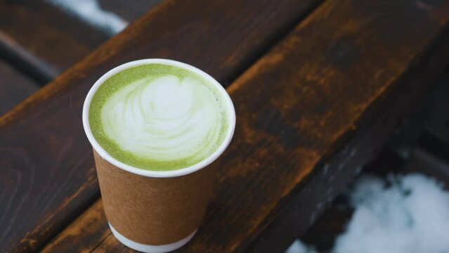 Paper cup with green matcha latte on wooden chair armrest.