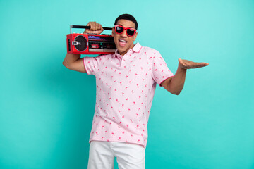 Portrait of cheerful carefree young man hold boombox enjoy music dancing isolated on turquoise...