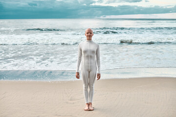 Full length portrait of young hairless girl with alopecia in white futuristic suit standing on sea...