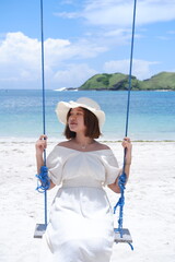 Woman in white dress and hat swinging on tropical beach, sunny day, good weather on lombok beach