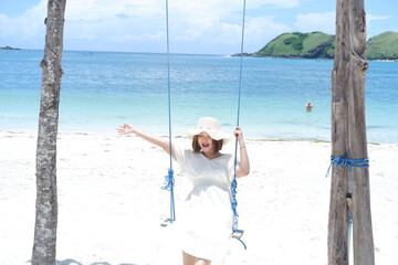 Woman in white dress and hat swinging on tropical beach, sunny day, good weather on lombok beach