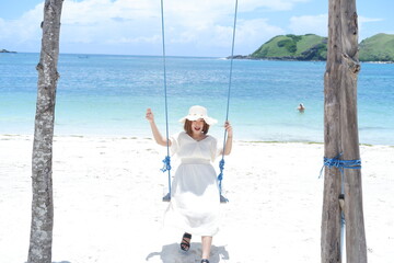 Woman in white dress and hat swinging on tropical beach, sunny day, good weather on lombok beach