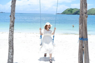 Woman in white dress and hat swinging on tropical beach, sunny day, good weather on lombok beach