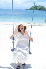 Woman in white dress and hat swinging on tropical beach, sunny day, good weather on lombok beach