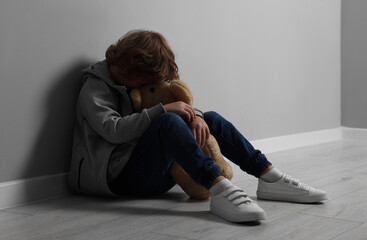 Child abuse. Upset boy with toy sitting on floor near grey wall