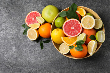 Different fresh citrus fruits and leaves in bowl on grey textured table, flat lay. Space for text