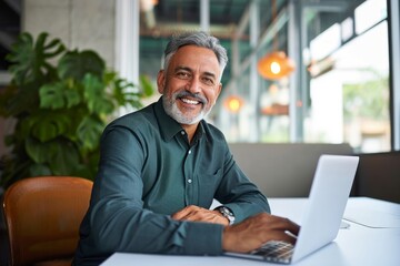 Happy smiling Indian older business man, middle aged professional businessman ceo executive bank manager, mature investor sitting at work desk in office with laptop looking at camera, Generative AI