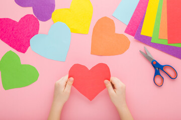 Little child hands holding red heart shape from application paper on light pink table background....