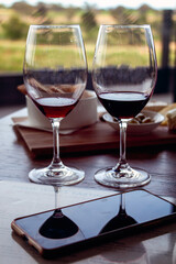 Two glasses of red wine and delicatessen food on wooden table. Platter of antipasto with cheese, crackers and olives. Glasses reflected on the screen of a cell phone. Background vineyards