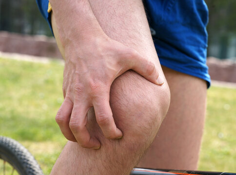Young man in sportswear on bike in urban park touching his painful knee
