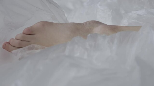 Female body part detail on the bed in the studio. Young woman feet close up seminude laying under thin sheet of oilcloth wrap.