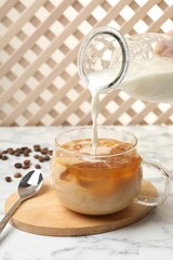 Woman pouring milk into glass with refreshing iced coffee at white marble table, closeup