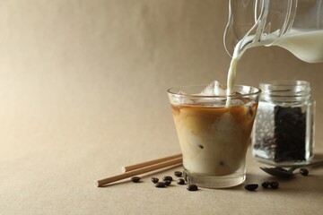 Pouring milk into glass with refreshing iced coffee on beige background, closeup. Space for text