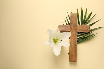 Wooden cross, lily flower and palm leaf on pale yellow background, top view with space for text. Easter attributes