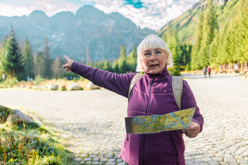 60s senior woman traveler with beige backpack with a paper map stands in a Tatra National Park, Poland. Adventurous woman navigating in with a topographic map in the beautiful polish mountains. 