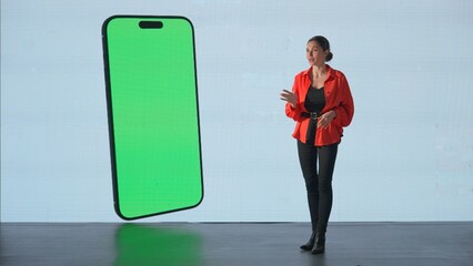 Woman presenting project on stage during meeting. Ceo businesswoman speaker presents product, smartphone chroma key green screen showing on on a huge LED screen.