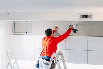 Plasterboard worker installs a plasterboard wall on the kitchen cabinets to cover the extractor pipe of the hood. - 742855186