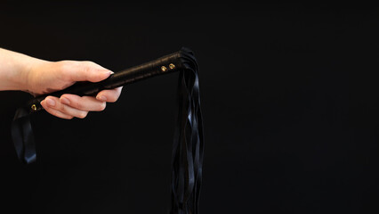 A woman's hand holds a whip against a dark background. Leather whip for spanking isolated on black....