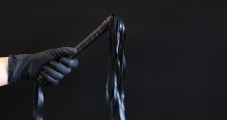 A hand in a black latex glove holds a whip against a dark background. Leather whip for spanking isolated on black. Sex toy for intimacy. Sexual slavery