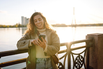 Young woman in coat enjoying views on embankment. Stylish woman with phone. Concept of vacation, technology, weekend.