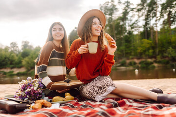 Two young women making picnic at the beach. Positive models sitting on a red mat with a thermos and...
