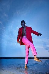 Fototapeta na wymiar stylish young african american man in pink pants and red blazer looking away on watery backdrop